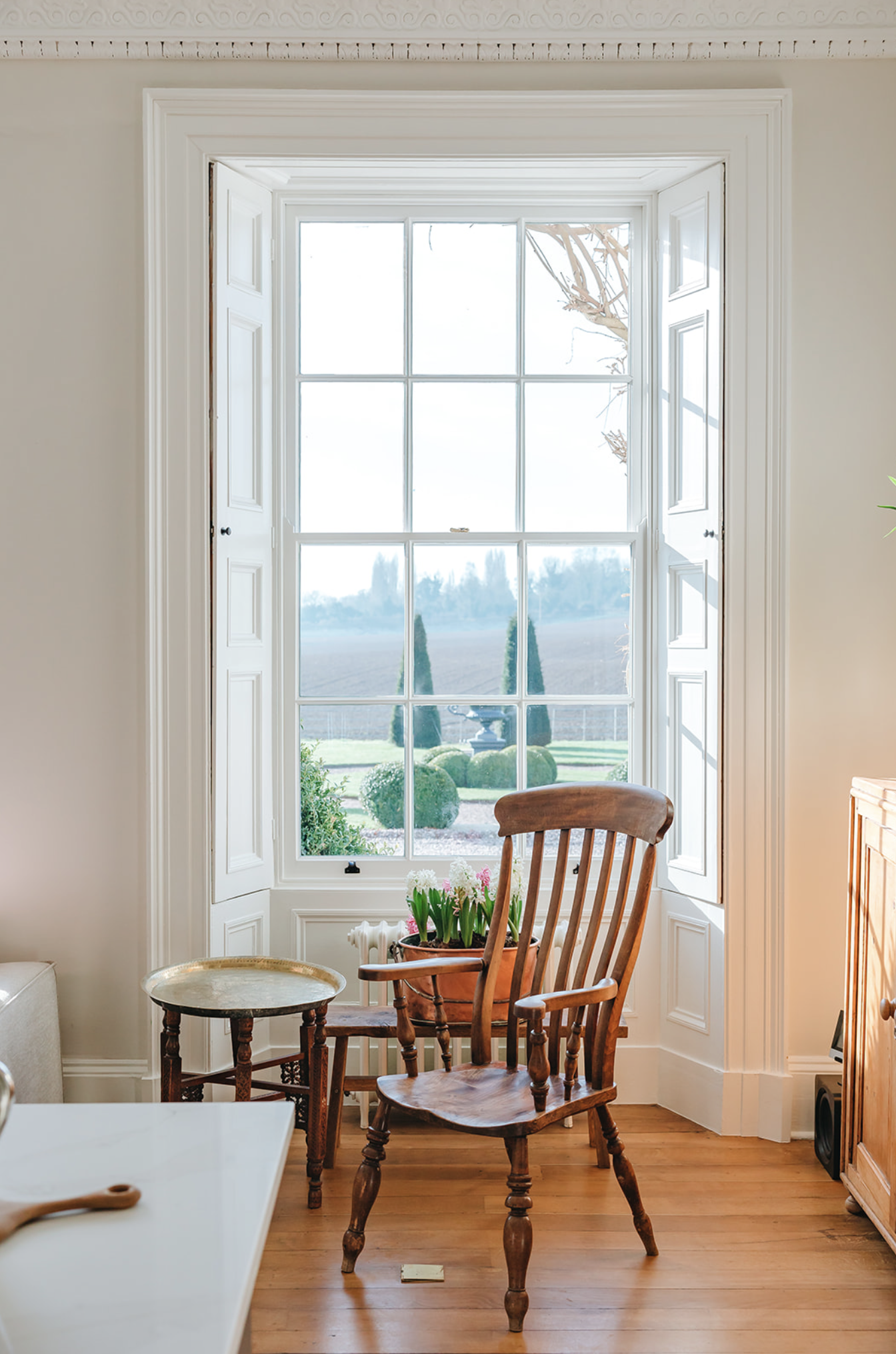 Tall sash window with working panelled shutters looking out over clipped topiary and open countryside, with a Windsor chair in front