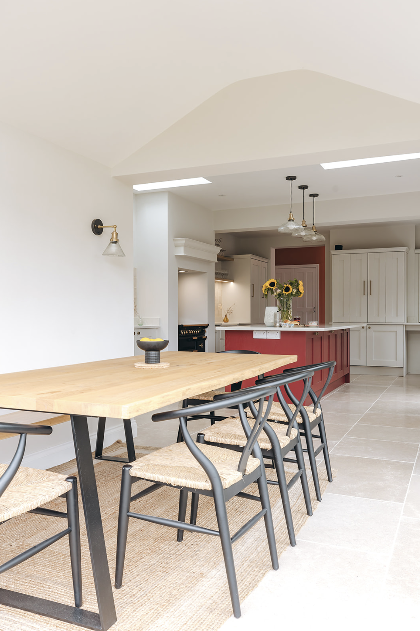Dining table with wishbone chairs beneath a vaulted ceiling and rooflight, looking through to the red-island kitchen and black range beyond