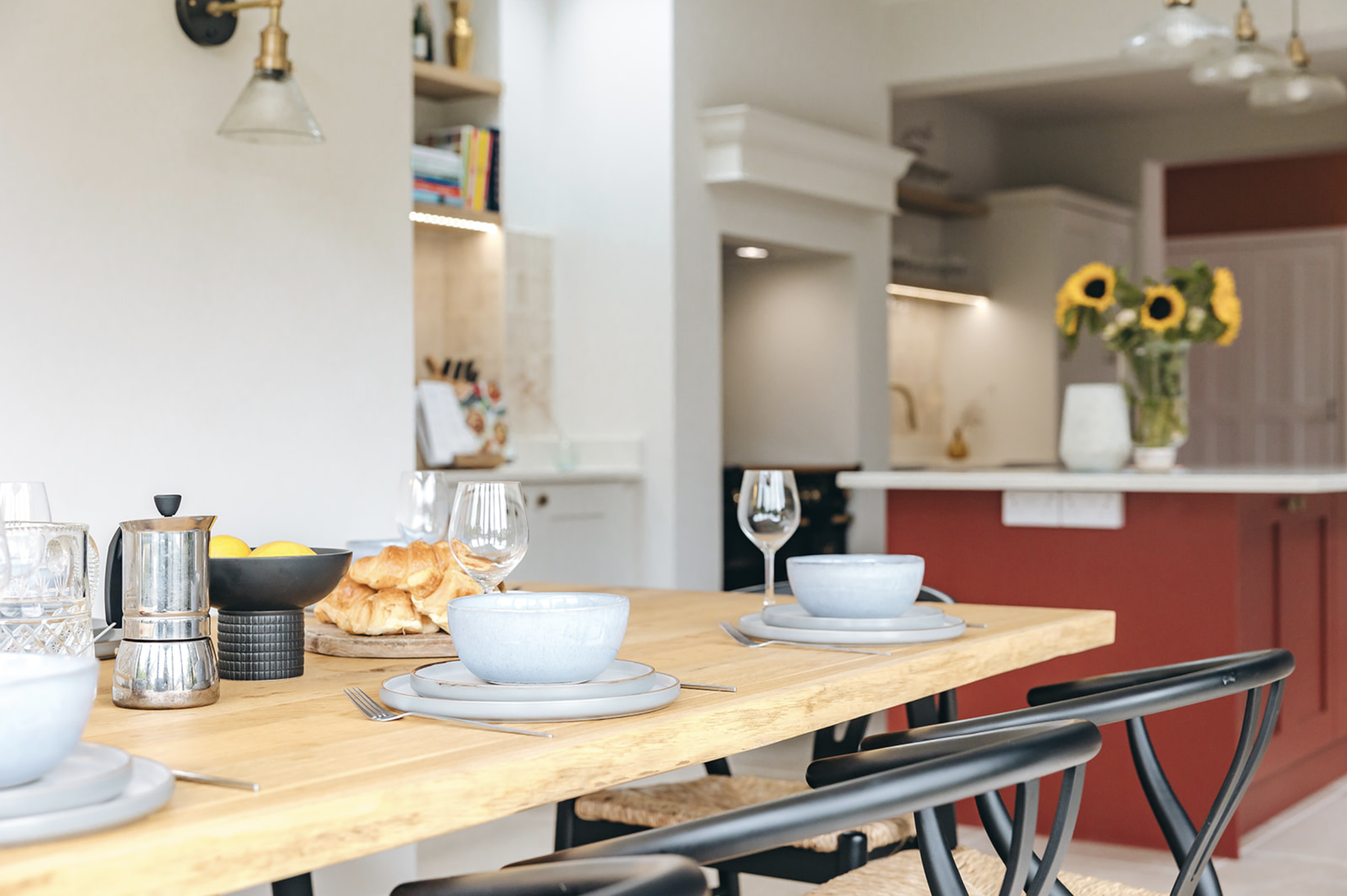 Oak dining table laid for breakfast, looking through to a kitchen with a deep red island and sunflowers