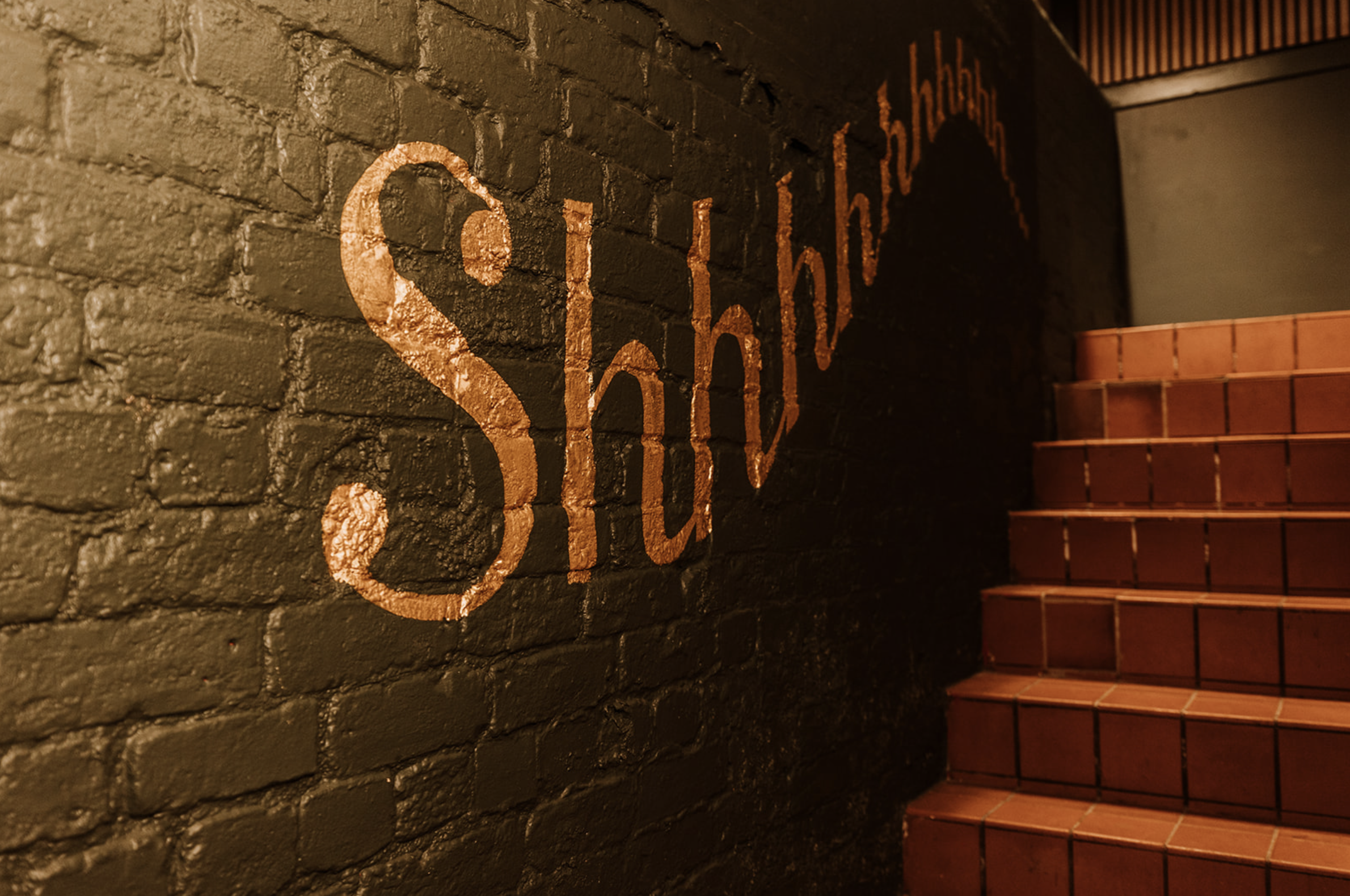 Gold-leaf Shhh signage on dark painted brick beside a tiled stair
