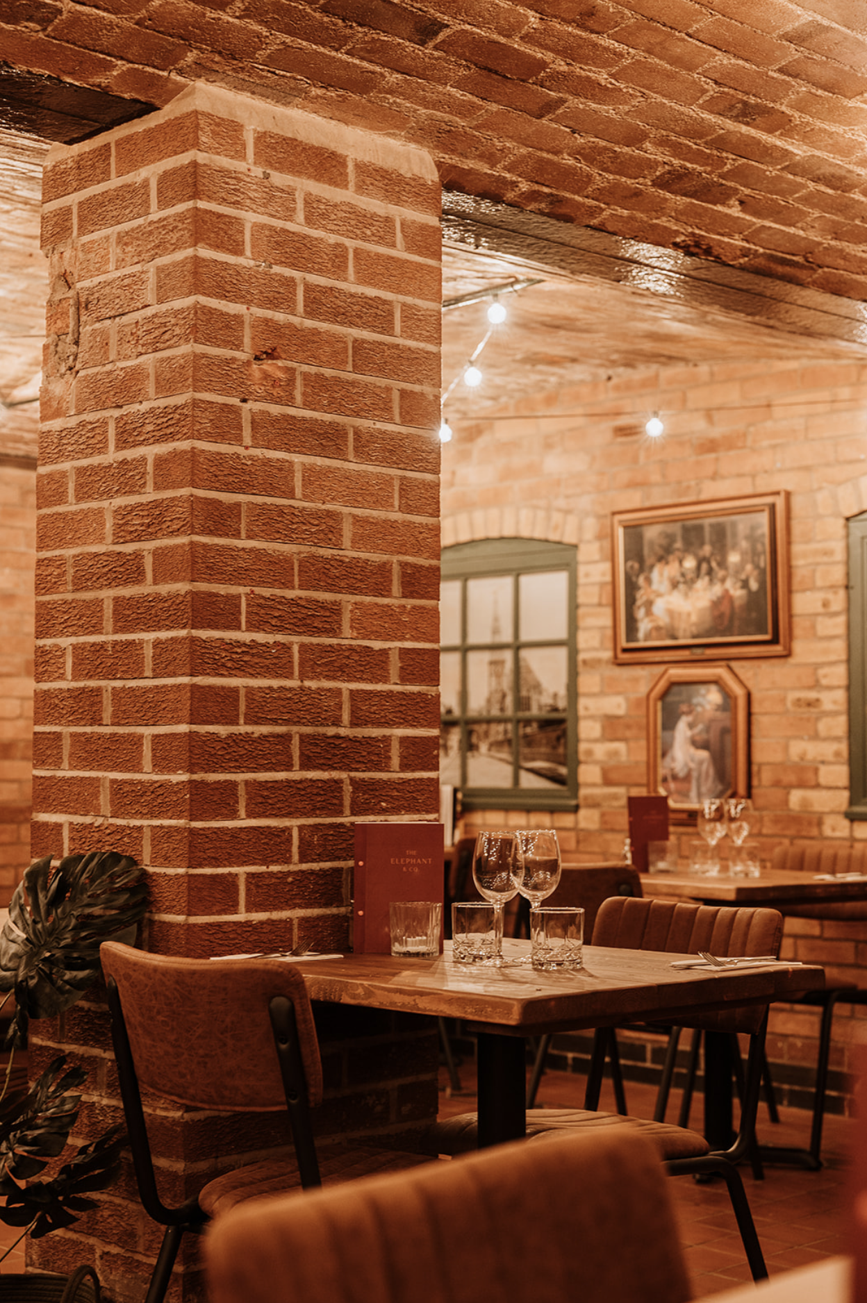 Dining table beside an exposed brick pillar hung with gilt-framed pictures