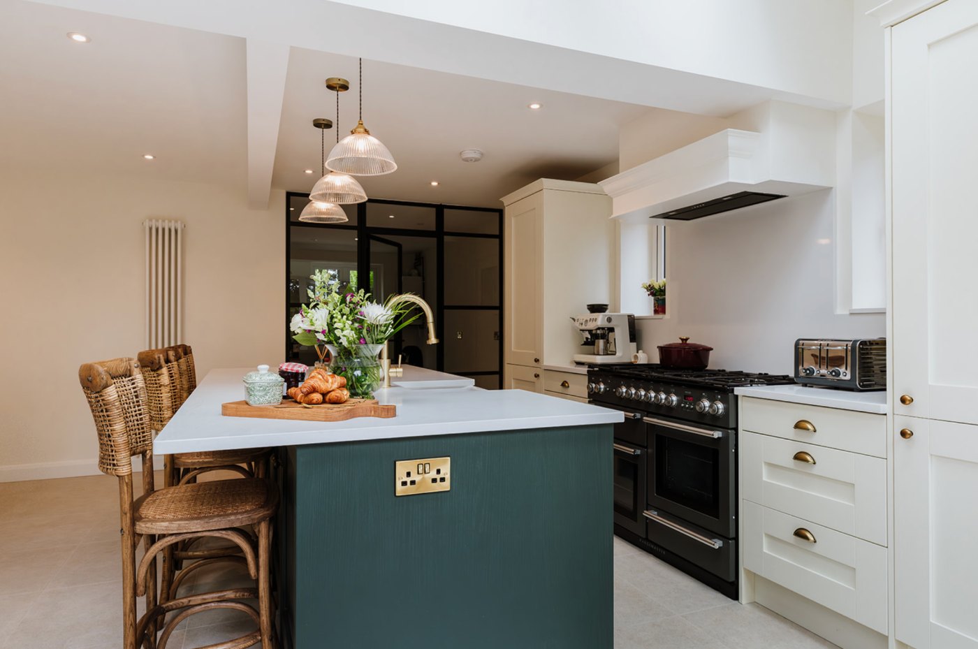 Kitchen with a dark teal island and white worktop, ribbed glass pendants, a black range set beneath a shaker canopy and a Crittall screen to the room beyond