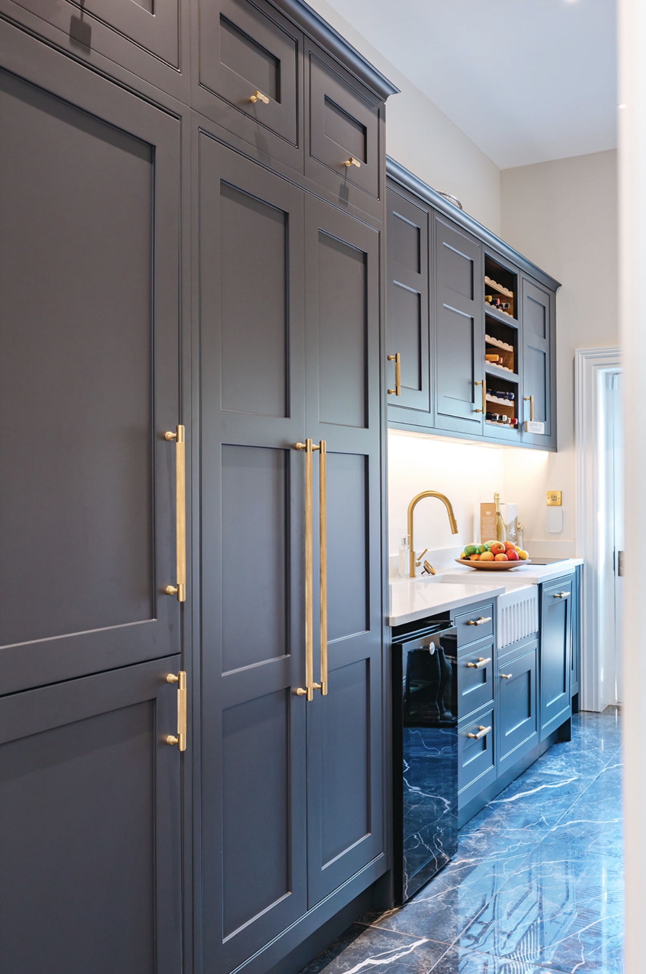 Long scullery with dark teal cabinetry, brass handles, white worktop and polished veined floor