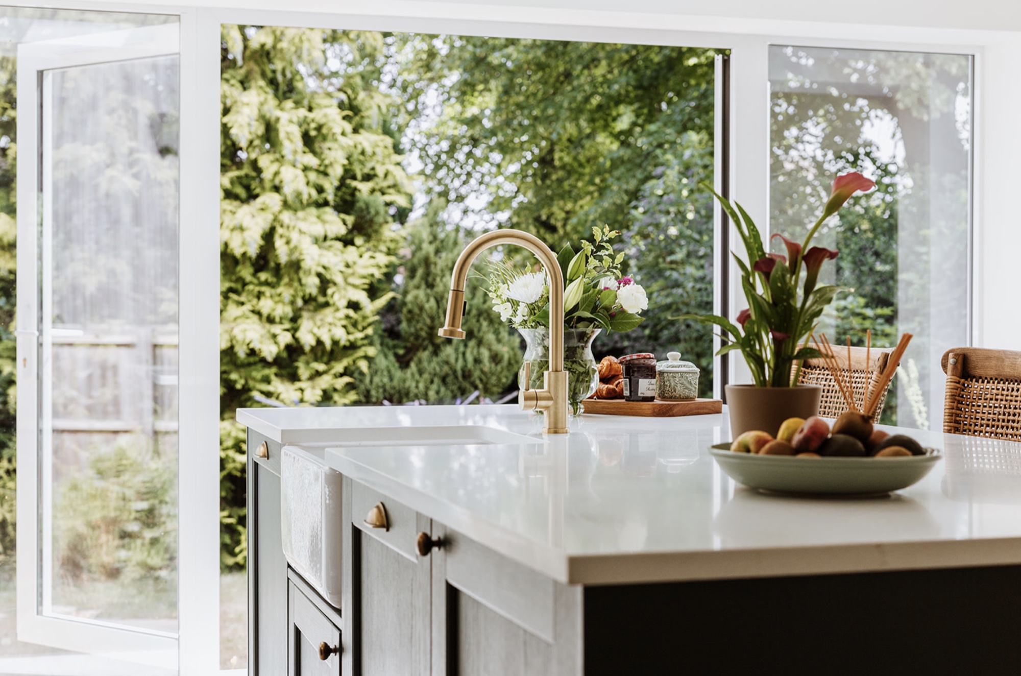 Long white kitchen island with brass tap, flowers and open doors to the garden