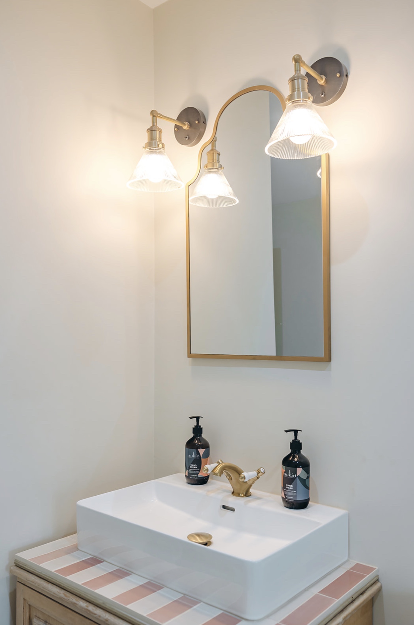 White cloakroom basin on a pink striped tiled vanity with brass tap, arched mirror and ribbed glass wall lights